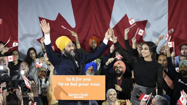 Jagmeet Singh en compagnie de sa conjointe et de militants du NPD qui agite des drapeaux du Canada sur une scène.