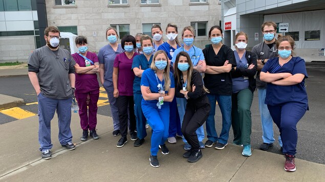 Une quinzaine de personnes  sont regroupés devant l'entrée d'un hôpital, pour la plupart les bras croisés. Ils portent un masque et sont habillés en tenue de personnel soignant.