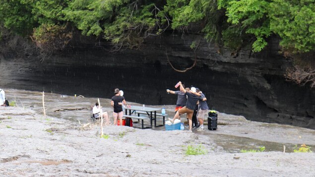 Plan large sur les abords d'une rivière où un groupe de personnes est attroupé dans une ambiance festive et décontractée. Certains esquissent quelques pas de danse joyeuse, d'autres s'enlacent.
