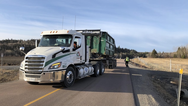 Un camion qui récupère les déchets au centre de la chaussée, près d'un agent de sécurité.