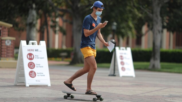 Un jeune homme qui porte des shorts jaunes et des sandales Birkenstock fait du skateboard avec un masque sur le visage, en regardant son téléphone en même temps.