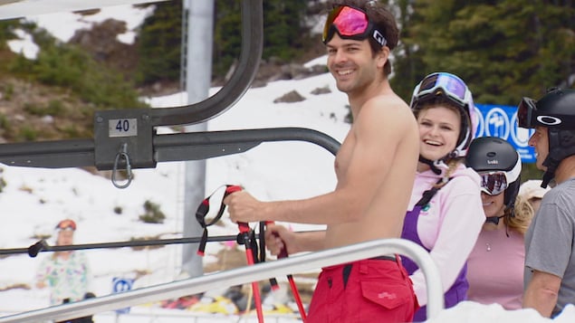 Un homme sans chandail avec des lunettes de ski sur la tête. Une femme est en arrière-plan.