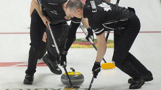 Brad Jacobs regarde Ryan Harnden et E.J. Harnden balayer sa pierre lors des essais olympiques de curling  à Saskatoon.