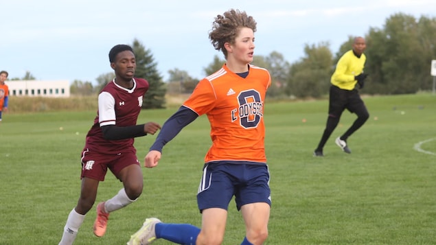 Un adolescent joueur de soccer habillé en maillot orange, courant sur le terrain cheveux au vent, un adversaire à ses trousses.