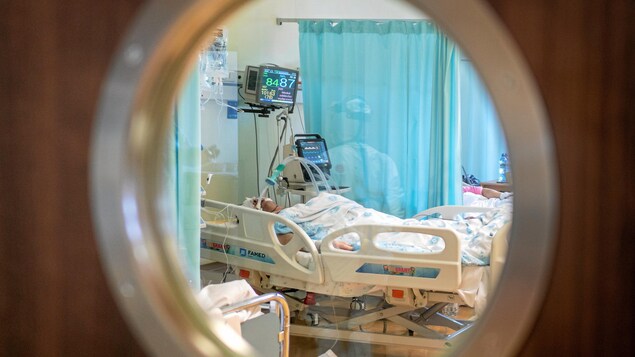 Une personne couchée dans un lit d'hôpital, photographiée à travers la fenêtre circulaire d'une porte fermée.