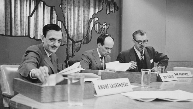 André Laurendeau, René Lévesque et Roger Duhamel consultant leurs notes sur un long bureau.