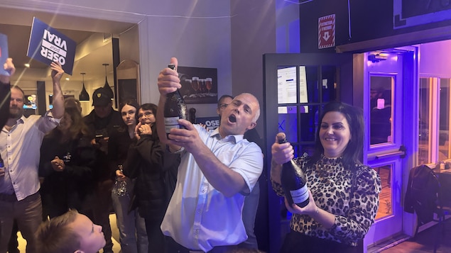 Il festoie et fait sauter le bouchon d'une bouteille de champagne.