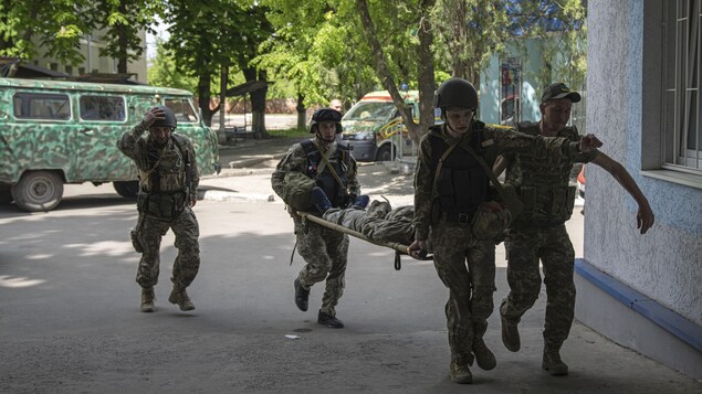 Trois soldats en transportent un autre sur une civière, près d'un bâtiment. 