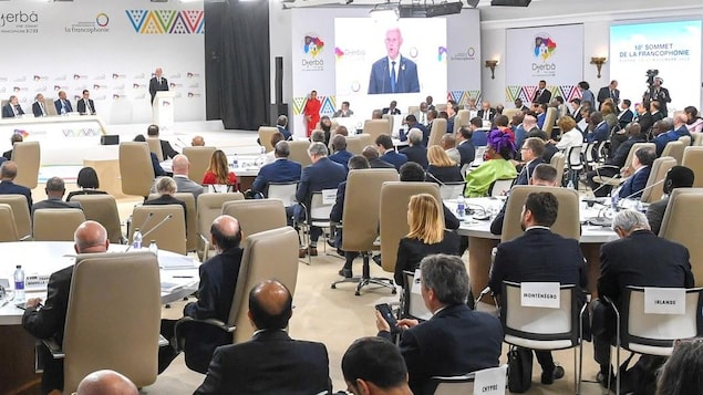 Une salle de conférence dans laquelle des dizaines de personnes sont assises sur des chaises face à une estrade et à de grands écrans. Sur les écrans, on voit le président de la Tunisie qui prononce une allocution.
