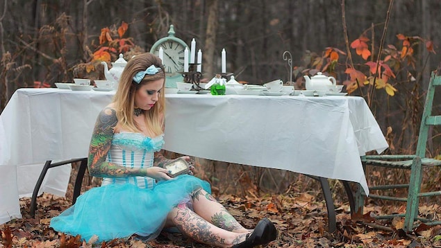 Une jeune femme porte une robe corset bleue claire à l'image d'Alice aux pays des merveilles. Elle est assise par terre. Ses bras et ses jambes sont tatoués. Le sol est recouvert de feuilles mortes. Une jeune femme porte une robe corset bleue claire à l'image d'Alice aux pays des merveilles. Elle est assise par terre. Ses bras et ses jambes sont tatoués. Le sol est recouvert de feuilles mortes.