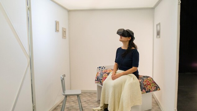 Une femme assise sur un petit lit assiste à un spectacle en réalité virtuelle au moyen de lunettes électroniques.