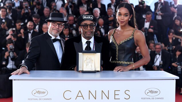 Le réalisateur Spike Lee tient le Grand Prix du Festival de Cannes sur un podium, entouré des acteurs Gary Oldman et Laura Harrier, avec toute une foule de photographes derrière lui. Le réalisateur Spike Lee tient le Grand Prix du Festival de Cannes sur un podium, entouré des acteurs Gary Oldman et Laura Harrier, avec toute une foule de photographes derrière lui.