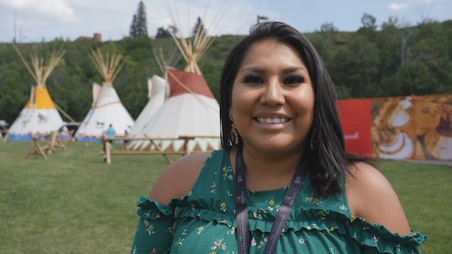 Une personne sourit à la caméra. Derrière elle, nous voyons des tipis.