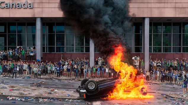 Una multitud frente a un edificio mira un vehículo volcado que está en llamas.