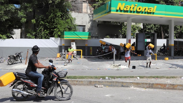 A man on a motorcycle drives by a petrol station while people carry gas cans on their shoulder.