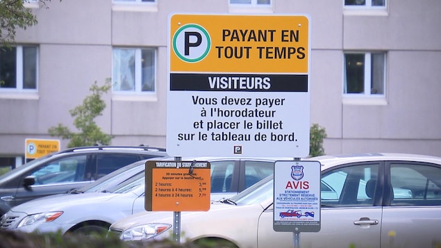 Dans le stationnement d'un hôpital, où se trouvent des automobiles, une pancarte indique : « Stationnement payant en tout temps ».