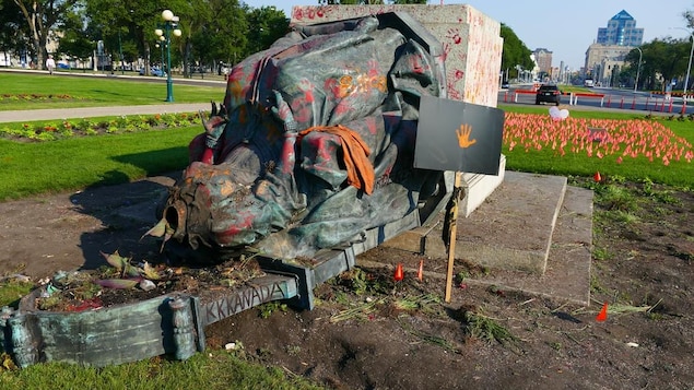 La statue, sans tête, est érendue par terre. Des drapeaux plantés dans le sol rappellent la mémoire des enfants autochtones morts dans les pentionnats.
