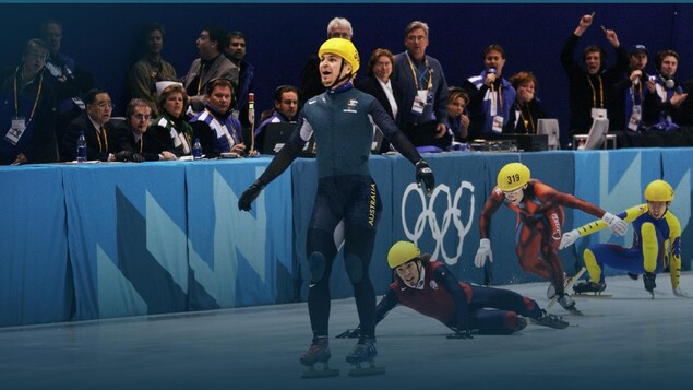 Un patineur de vitesse passe la ligne d'arrivée sous le regard de trois adversaires qui sont par terre sur la glace.