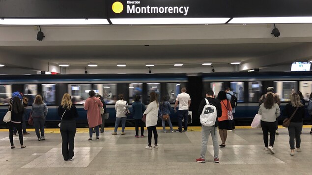 Une rame de métro passe, tandis que des usagers attendent sur le quai.