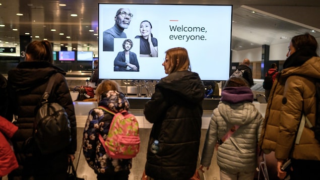 People are waiting for their suitcases in front of a conveyor belt. A screen displays "welcome everyone".