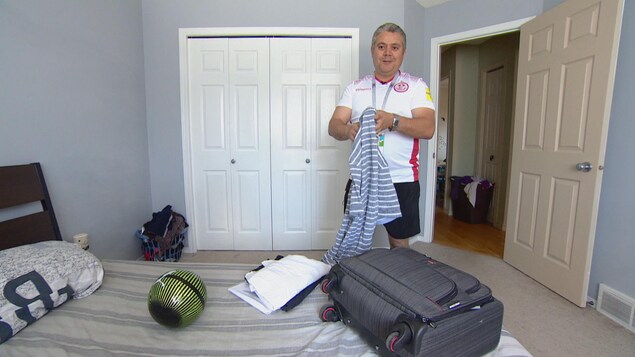 Un homme vêtu d'un maillot de soccer tient une chemise, devant une valise posée sur un lit, dans une chambre. Un homme vêtu d'un maillot de soccer tient une chemise, devant une valise posée sur un lit, dans une chambre.