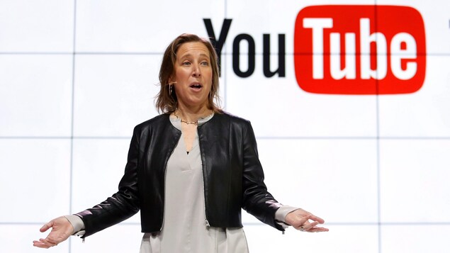 Susan Wojcicki est debout devant un écran où est affiché le logo de YouTube.