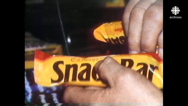 Main tenant une tablette de chocolat Snack Bar et pointant le nom de la marque Cadbury.