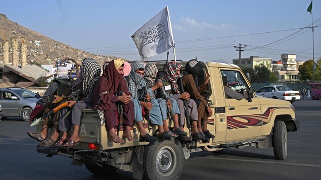 Plusieurs hommes armées sont assis dans la boîte d'une camionnette, surmontée d'un drapeau des talibans. 