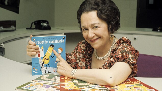Dans un bureau, Lucille Desparois présente un de ses livres de contes parmi ceux qui sont étalés sur la table devant elle. Dans un bureau, Lucille Desparois présente un de ses livres de contes parmi ceux qui sont étalés sur la table devant elle.