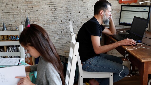 Une fillette fait ses devoirs, assise à une table, alors que son père travaille à l'ordinateur.
