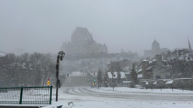 La silhouette emblématique de bâtiments comme le Château Frontenac et l'ancien bureau de poste de Québec se dessine dans un ciel gris pendant une tempête de neige.