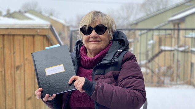 Une femme dehors, portant des lunettes de soleil et tenant dans ses mains un cahier noir.