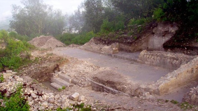 On voit une place rectangulaire excavée, entourée de murets de pierre, dans un site où la végétation est abondante.