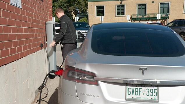 On voit l'arrière d'une voiture avec un fil qui la relie à une borne électrique. Un homme roule le fil. On voit l'arrière d'une voiture avec un fil qui la relie à une borne électrique. Un homme roule le fil.