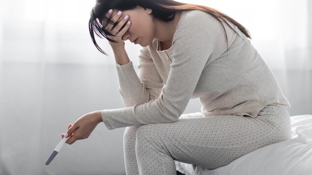 Une femme en pyjama assise sur le bord de son lit, en état de choc, le front appuyé dans une main et l'autre main tenant un test de grossesse fait à la maison.