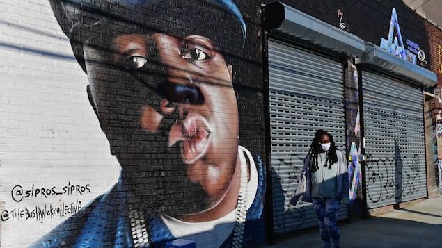 Une murale représentant le rappeur The Notorious B.I.G. dans une rue à New York, devant laquelle passe une piétonne portant un masque chirurgical. 