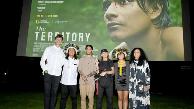 Six personnes posent côte à côte sur un parterre de pelouse, devant un écran géant présentant l'affiche du documentaire The Territory. 