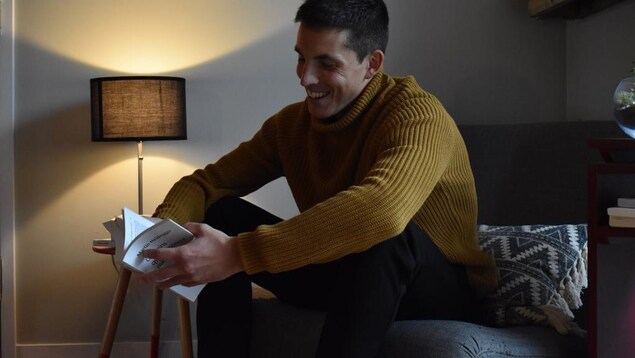 Portrait en couleur de Thomas Windish avec une lampe allumée en arrière plan. Le jeune homme, souriant, est assis sur un fauteuil gris et tient dans ses mains un livre de Heidegger.