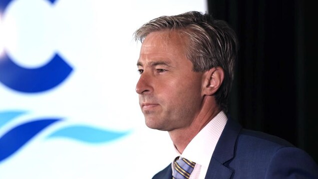 Tim Houston photographié de profil à un lutrin devant deux micros. Derrière lui se trouve un grand écran sur lequel apparaît le logo du Parti progressiste-conservateur de la Nouvelle-Écosse.