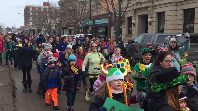 Des dizaines d'adultes et d'enfants dont certains tiennent de petits drapeaux fransaskois marchent dans une rue.