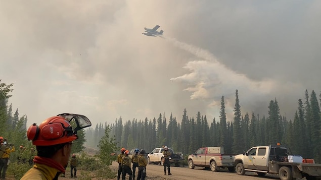 Avion-citerne qui déverse de l'eau sur la forêt pendant que des pompiers forestiers sont au sol et observent la scène, en juin 2024, à Fort Good Hope, aux Territoires du Nord-Ouest.