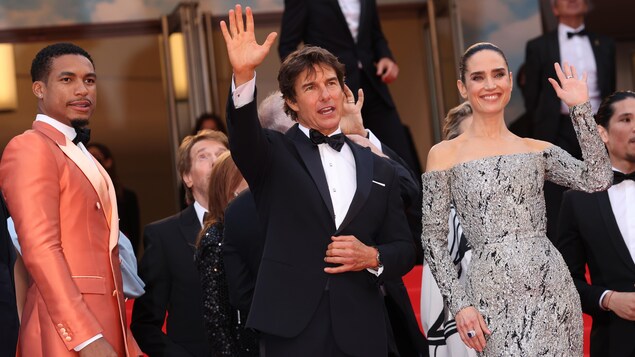 Un homme en smoking, accompagné d'un autre homme portant un costume orange et d'une femme en robe grise, salue la foule sur le tapis rouge du Festival de Cannes. 
