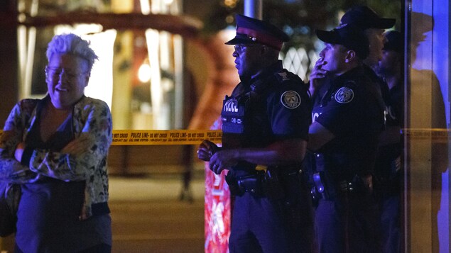 Des policiers en uniforme regardent droit devant eux, l'air penseur, alors qu'une femme aux cheveux courts pleure près d'eux. La photographie a été prise le soir ou la nuit, puisqu'il fait sombre. Des policiers en uniforme regardent droit devant eux, l'air penseur, alors qu'une femme aux cheveux courts pleure près d'eux. La photographie a été prise le soir ou la nuit, puisqu'il fait sombre.