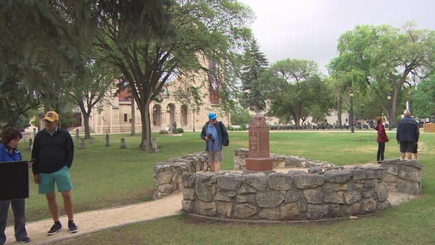 Trois personnes se situent à l'avant-plan, deux regardent un panneau indicatif de l'histoire de Louis Riel et le dernier tient un appareil photo à la main, à côté de la tombe. A l'arrière-plan à droite, à proximité de la tombe, trois autres personnes discutent. On voit la cathédrale de Saint-Boniface en arrière-plan. Trois personnes se situent à l'avant-plan, deux regardent un panneau indicatif de l'histoire de Louis Riel et le dernier tient un appareil photo à la main, à côté de la tombe. A l'arrière-plan à droite, à proximité de la tombe, trois autres personnes discutent. On voit la cathédrale de Saint-Boniface en arrière-plan.