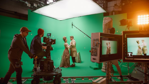 Une équipe tourne une scène de film dans un studio devant un écran vert, avec un acteur et une actrice habillés en costumes d'époque au centre. 