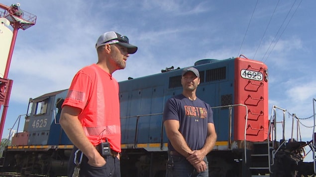 Deux hommes, à l'extérieur, devant une locomotive. Au fond à gauche, on voit un petit coin de la station de chargement du grain, devant le chemin de fer.
