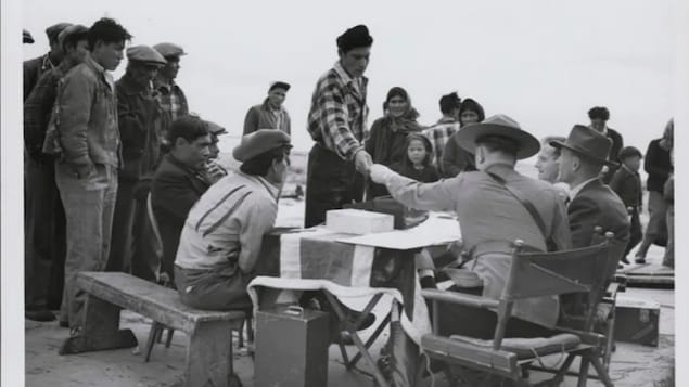 A Dene man accepts money from a fully uniformed Mountie seated behind a table draped with a British flag in Yellowknife. Food and money were distributed in communities after — and sometimes before — Treaty 11 was signed.