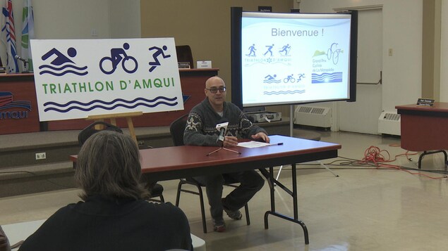 Homme assit à une table devant des panneaux : Triathlon d'Amqui Homme assit à une table devant des panneaux : Triathlon d'Amqui