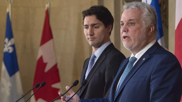 Un homme aux cheveux blancs et ayant une petite barbe blanche répond à une question. Un autre homme plus jeune se tient derrière lui. Ils sont entourés de drapeaux du Québec et du Canada,