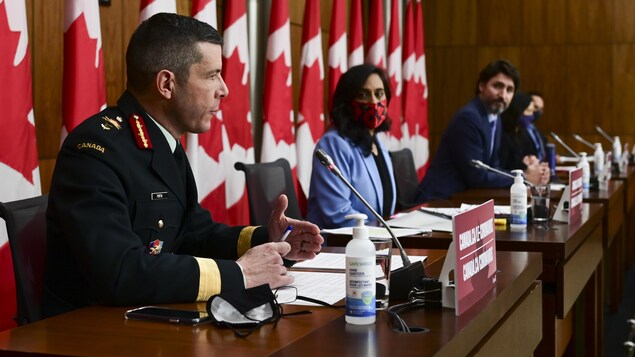 Des personnes assises à des bureaux derrière des micros et devant des drapeaux canadiens.